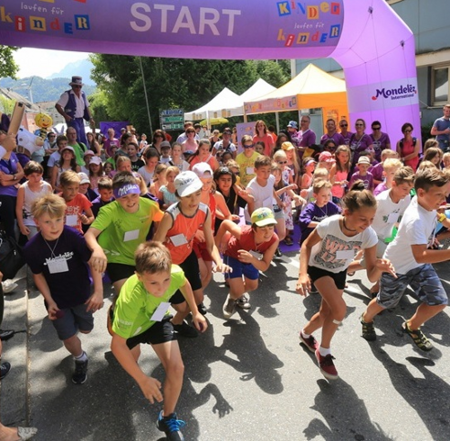 Viele Kinder rennen lachend beim Start eines Benefizlaufs los. Ein lilafarbenes Tor mit der Aufschrift „Kinder laufen für Kinder“ spannt sich über der Straße. Dahinter jubeln Eltern und Zuschauer:innen.